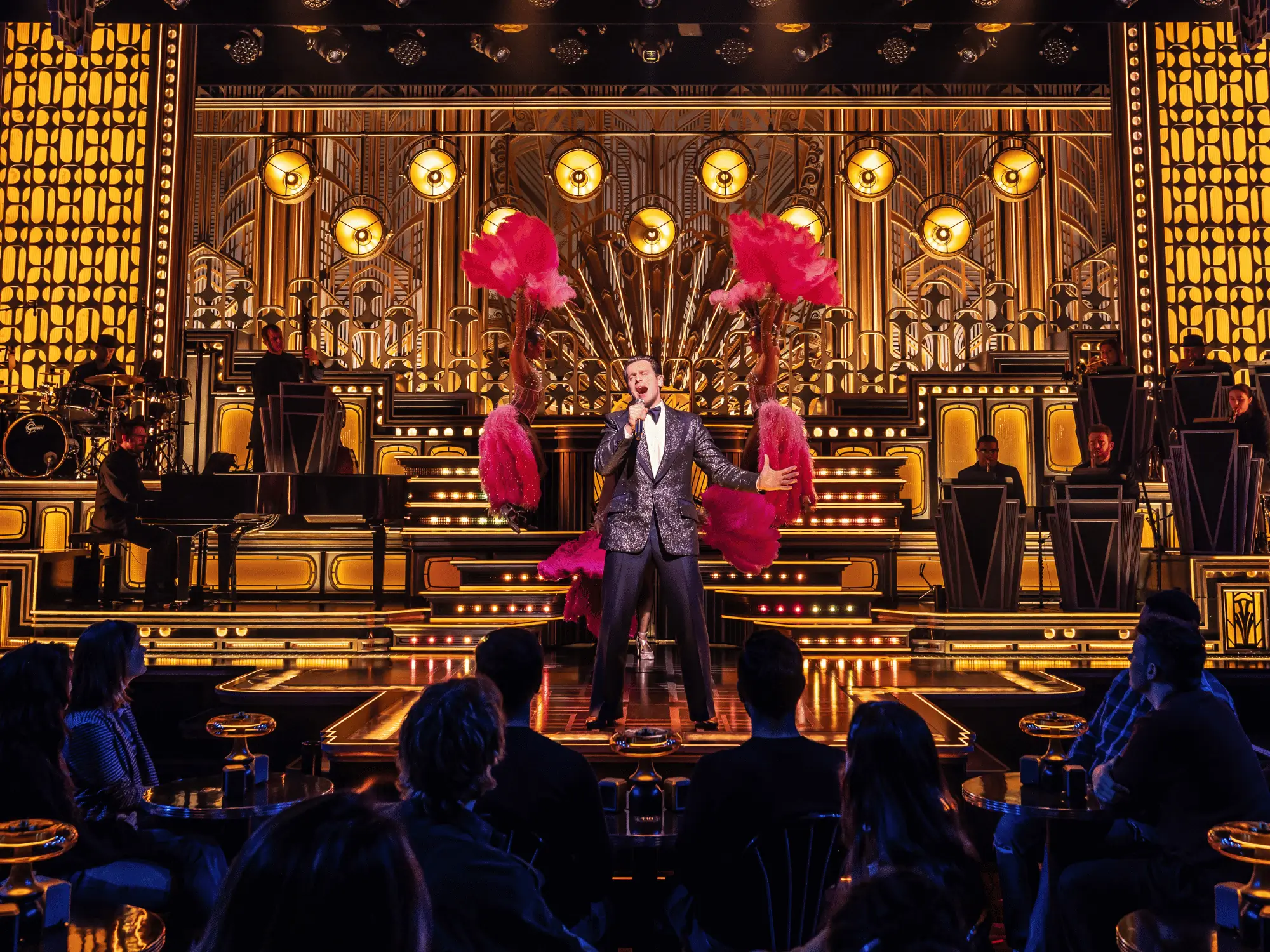 A singer in a sparkling tuxedo performing center stage on a glittering Art Deco nightclub set, flanked by dancers with pink feather fans