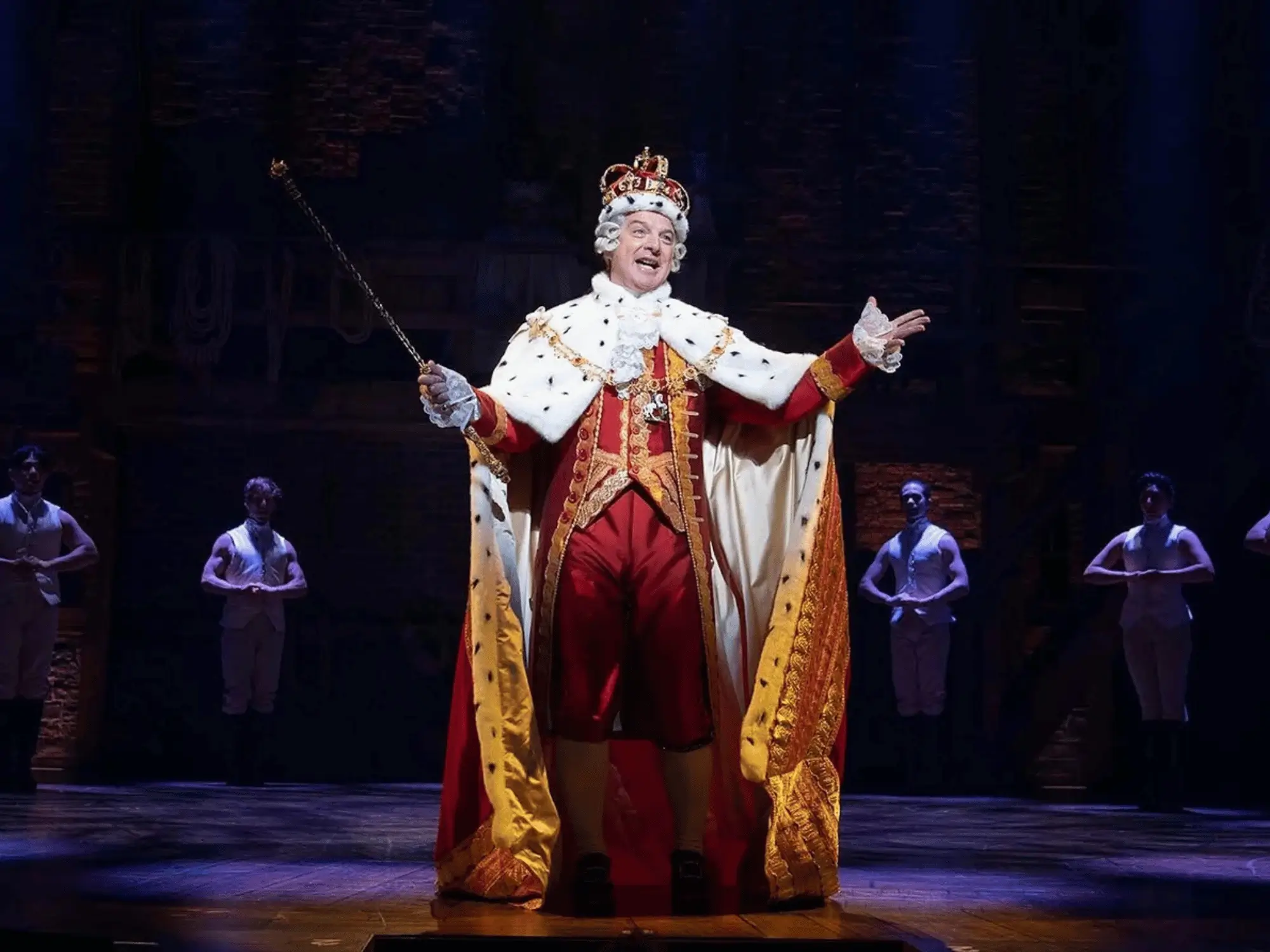 An actor as King George wearing a jeweled crown and ermine robe, arms outstretched on a Broadway stage