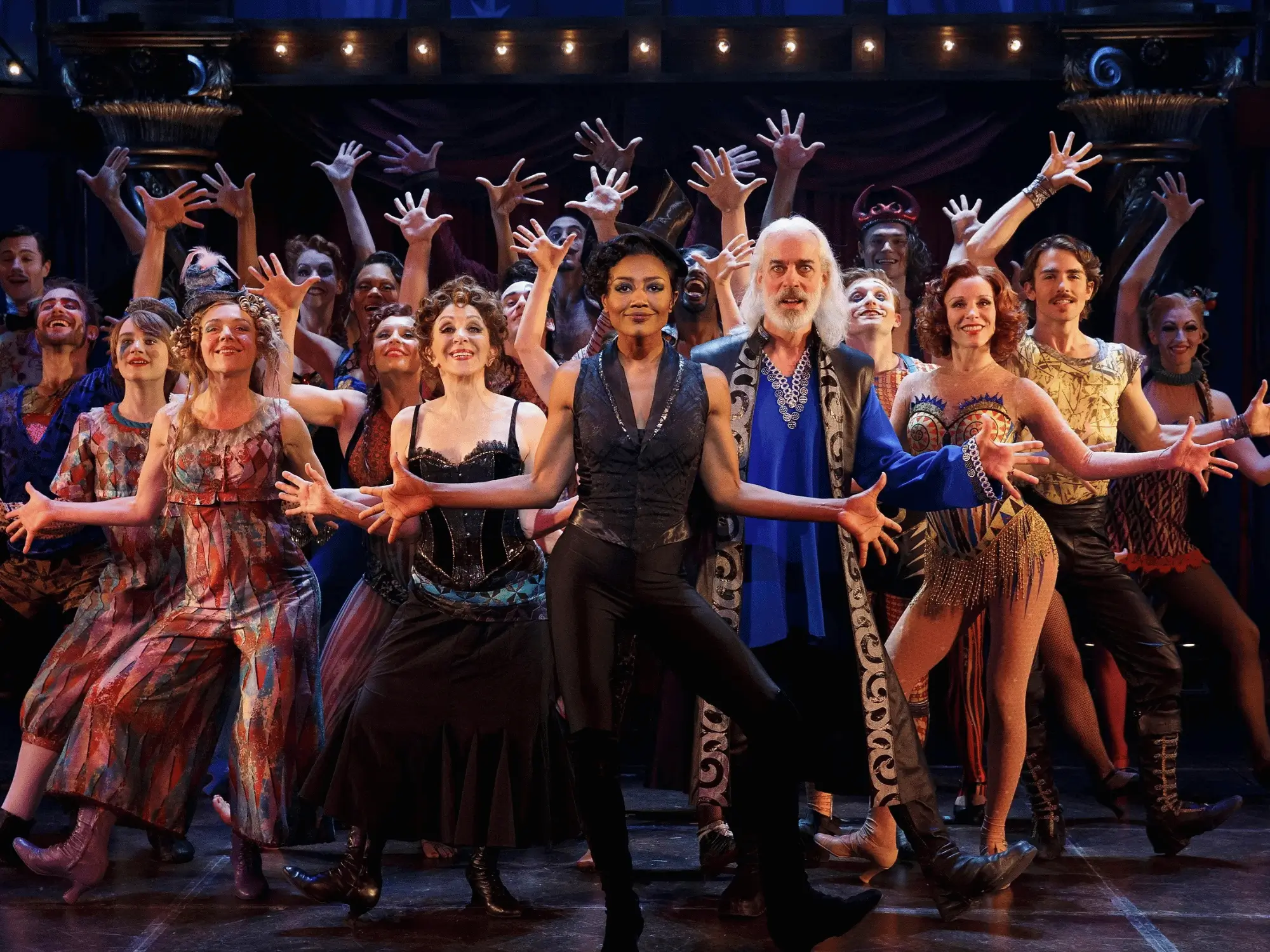 A Broadway ensemble in vibrant costumes dancing with arms raised during a musical number
